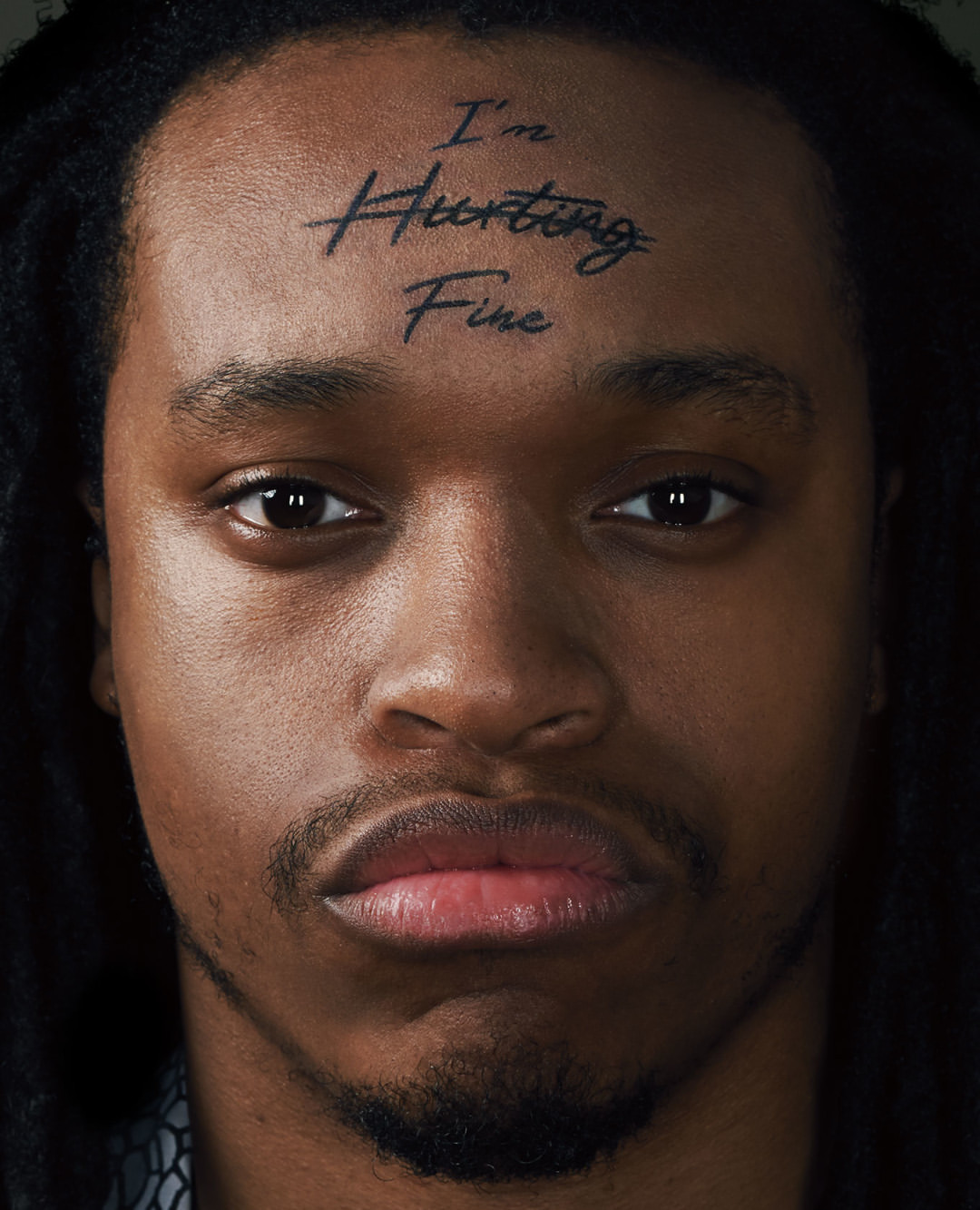 Close-up portrait of a Black adult male with dark dreadlocks, a serious facial expression, and the words 'I'm Hurting Fine' tattooed on his forehead.