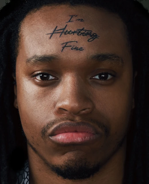 Close-up portrait of a Black adult male with dark dreadlocks, a serious facial expression, and the words 'I'm Hurting Fine' tattooed on his forehead.