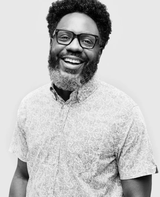 Portrait of an adult male with dark hair and a grey beard, wearing glasses and a patterned shirt, smiling against a neutral background.