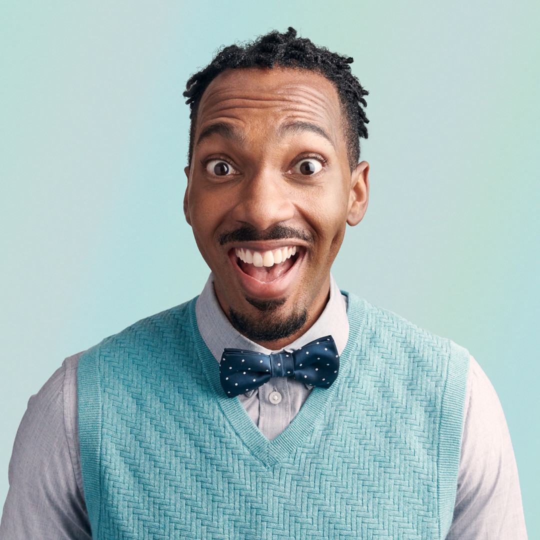 Close-up portrait of a smiling African-American man with a bow tie, wearing a sweater vest in a photo studio with light blue background.