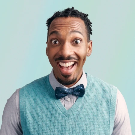 Close-up portrait of a smiling African-American man with a bow tie, wearing a sweater vest in a photo studio with light blue background.