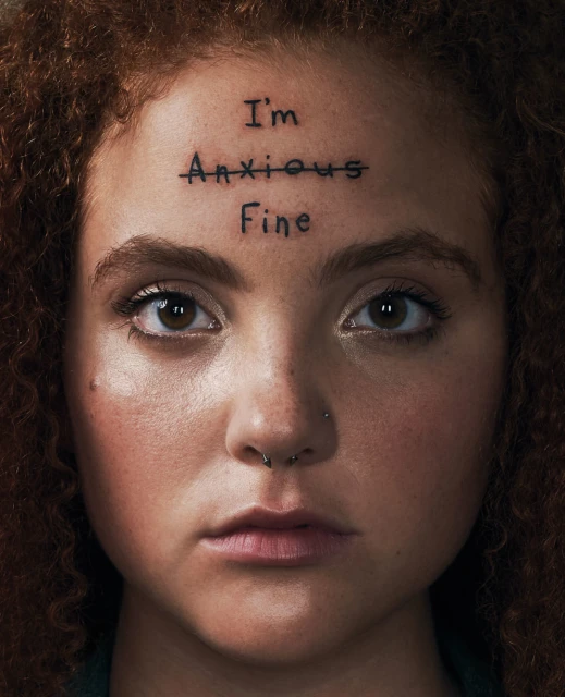 Close-up of a young adult woman with curly red hair and light skin, looking directly at the camera with text on her forehead.