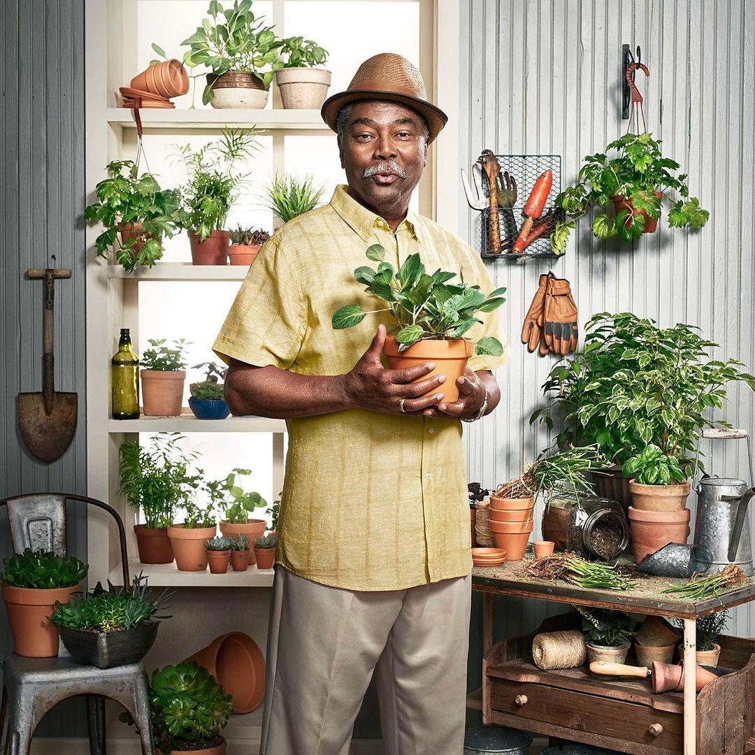 A smiling older man with a hat holds a potted plant inside a plant-filled indoor potting area, surrounded by terracotta pots and greenery.