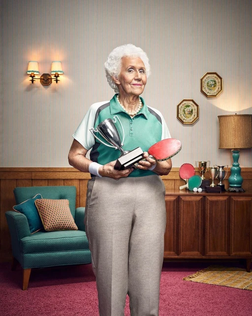 Elderly woman smiles, holding a trophy and ping pong paddle in an indoor setting with vintage furniture and decor.