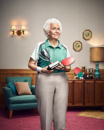 Elderly woman smiles, holding a trophy and ping pong paddle in an indoor setting with vintage furniture and decor.