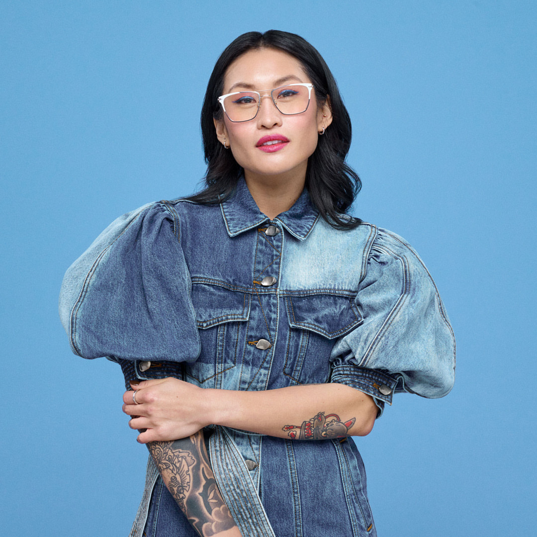 Portrait of an Asian woman wearing eyeglasses and a two-tone denim jacket against a blue background in a studio setting.