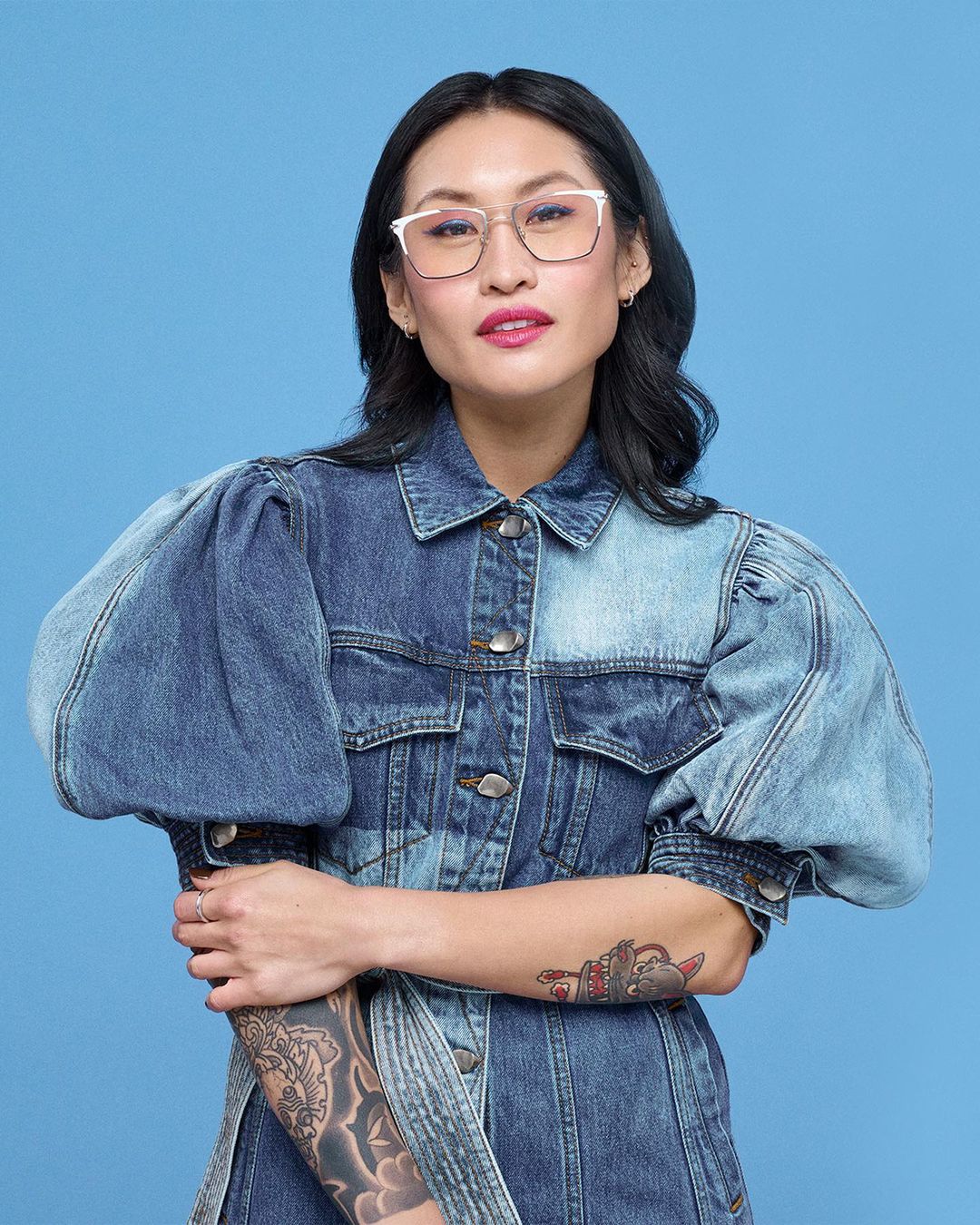 Woman wearing glasses and a denim jacket posing against a blue background, showcasing fashion and portrait photography.