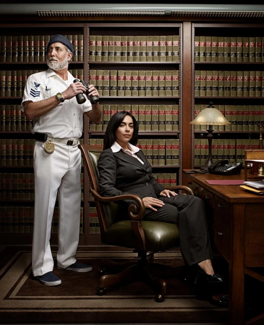 Indoor portrait of an adult man wearing a captain's uniform with binoculars stands next to an adult woman in business suit in office setting.