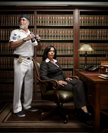 Indoor portrait of an adult man wearing a captain's uniform with binoculars stands next to an adult woman in business suit in office setting.