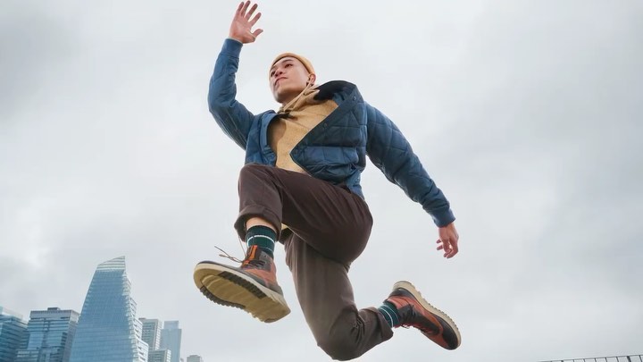 Young adult in mid-air wearing a blue puffer jacket, tan hoodie, brown pants, and hiking boots, city buildings and cloudy sky background.