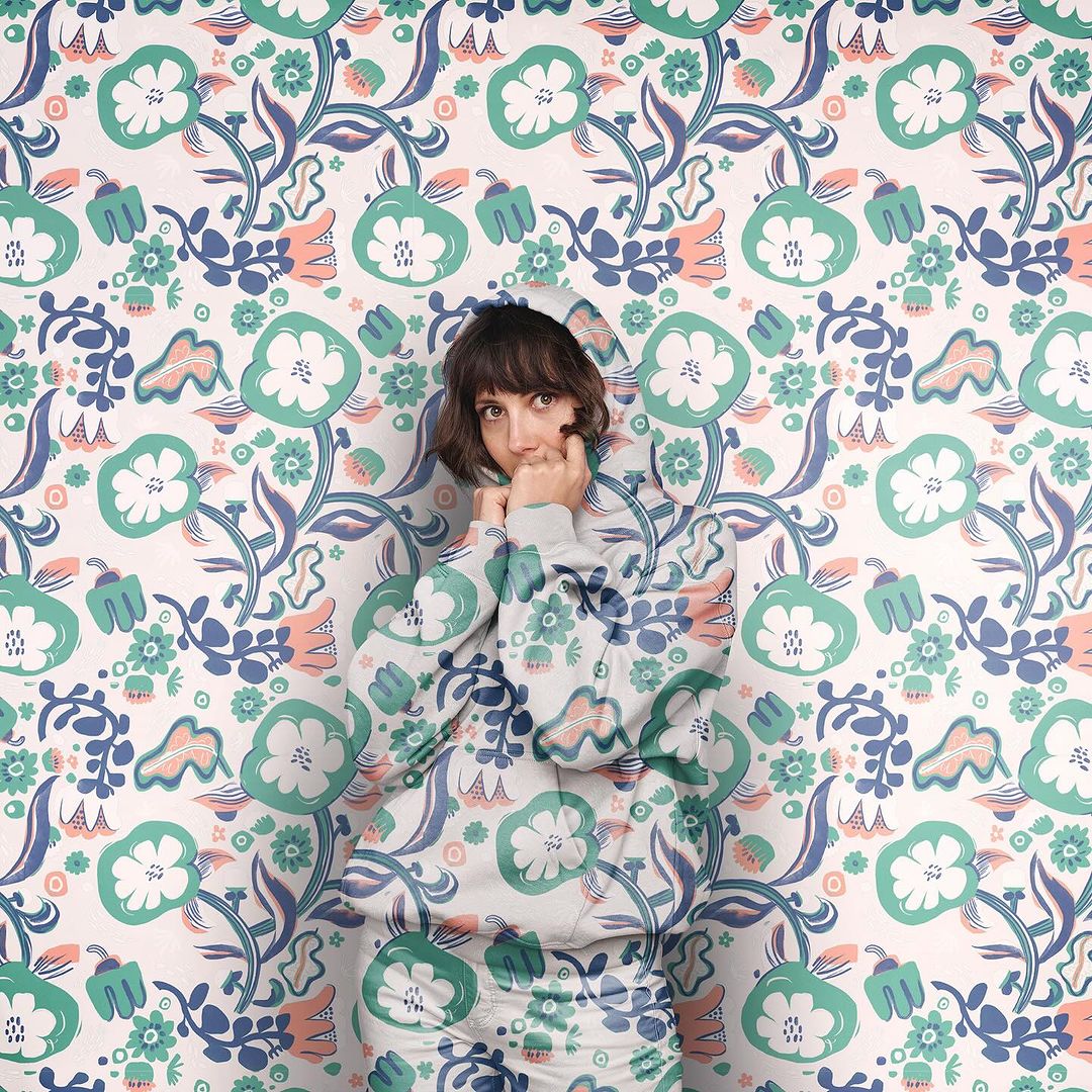 Adult woman in floral-patterned hoodie blends into matching floral wallpaper. Portrait with direct gaze, fair skin, and short brown hair.