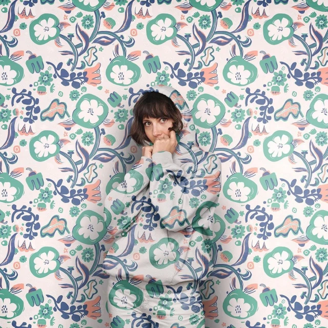 Adult woman in floral-patterned hoodie blends into matching floral wallpaper. Portrait with direct gaze, fair skin, and short brown hair.