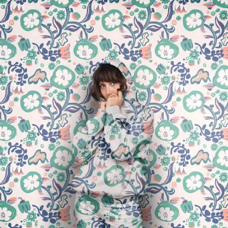 Adult woman in floral-patterned hoodie blends into matching floral wallpaper. Portrait with direct gaze, fair skin, and short brown hair.