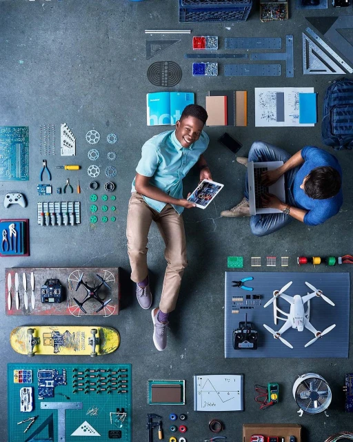 Overhead shot of two men sitting on a concrete floor surrounded by electronics, circuit boards, drone, tools, skateboard, and laptop.