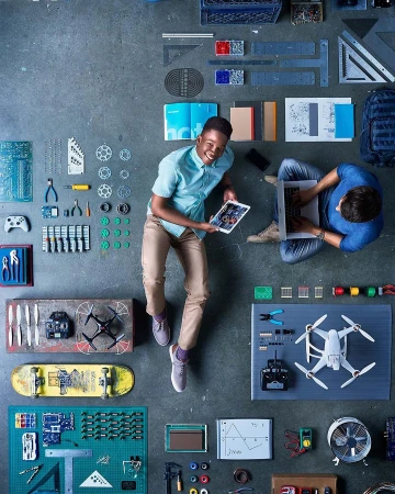 Overhead shot of two men sitting on a concrete floor surrounded by electronics, circuit boards, drone, tools, skateboard, and laptop.