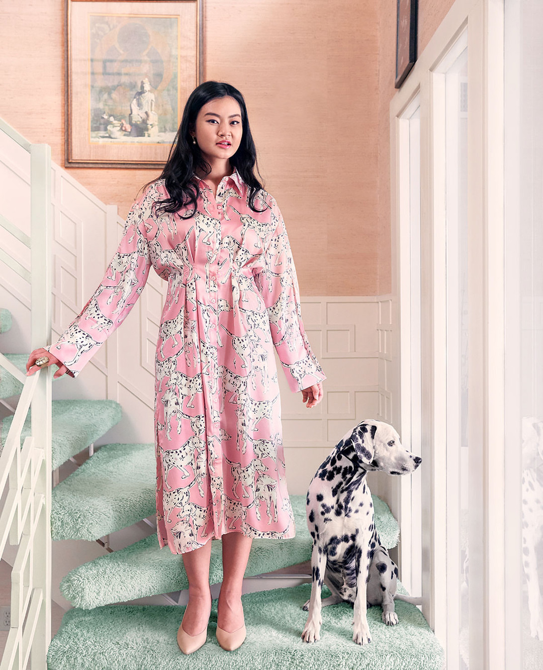 An Asian woman stands on a carpeted staircase in a pink Dalmatian-print dress next to a Dalmatian dog indoors with natural light.