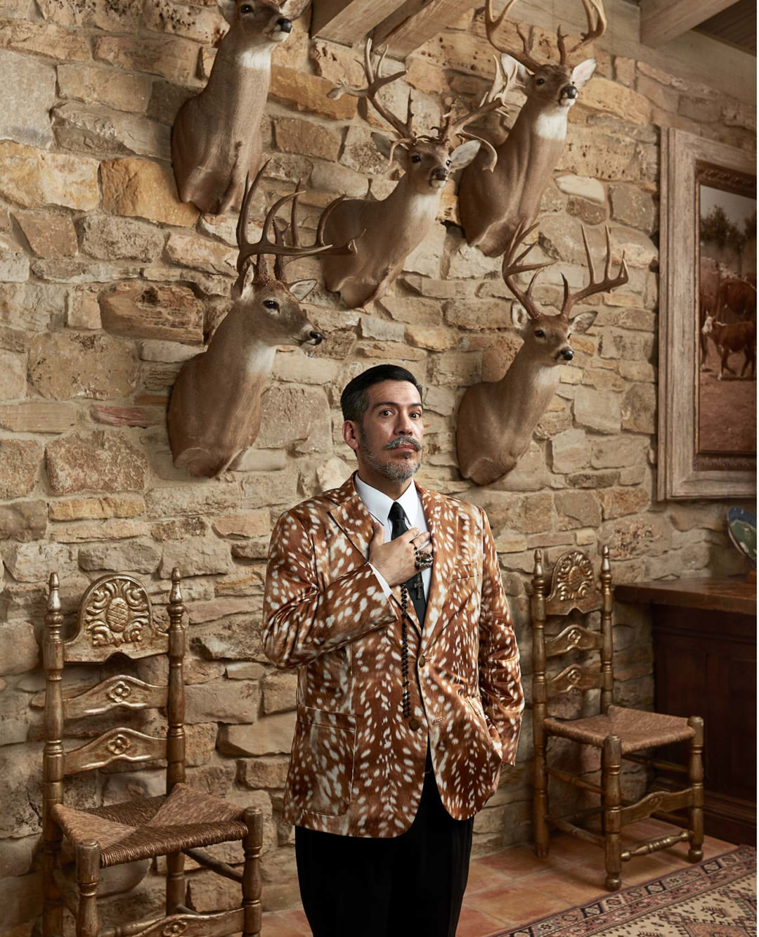 Stylish man in a patterned jacket poses in an ornate room with mounted deer heads on a stone wall, seated ornate chairs.