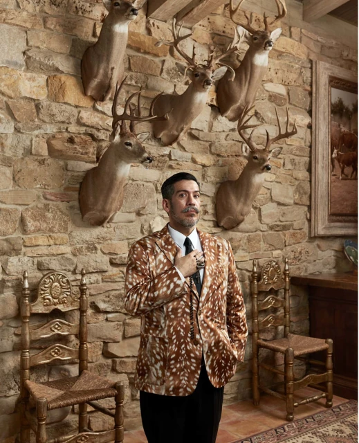 Stylish man in a patterned jacket poses in an ornate room with mounted deer heads on a stone wall, seated ornate chairs.
