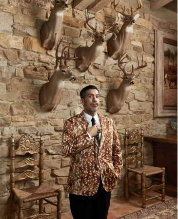 Stylish man in a patterned jacket poses in an ornate room with mounted deer heads on a stone wall, seated ornate chairs.