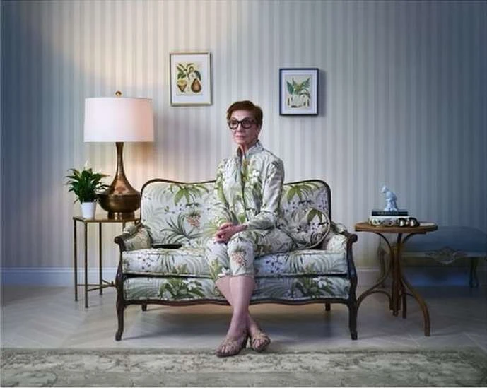 Elegant woman in floral suit sits on a sofa in a well-lit room, complemented by side tables, wall art, and a striped wallpaper backdrop.