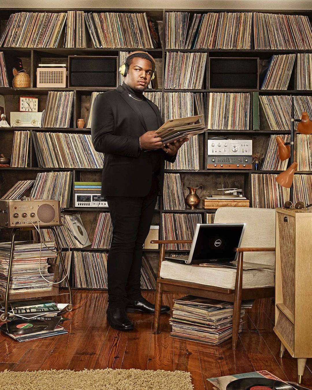 Man in a suit holding vinyl records stands in front of a large record collection with a laptop, radio, and vintage stereo system.