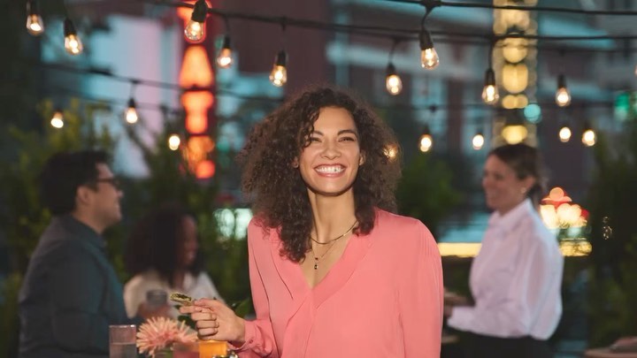 Smiling woman poses outdoors in warm light, enjoying an outdoor event with friends at dusk under strings of lights in an urban setting.