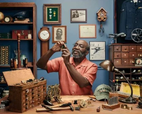 A person wearing a collared shirt and glasses sits at a desk, meticulously repairing a watch with tools in a room with memorabilia.