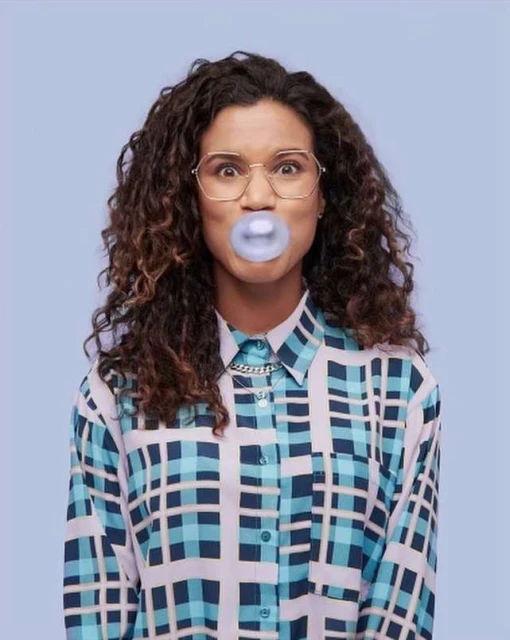 Adult female with curly brown hair, wearing glasses and a turquoise patterned shirt, blowing a bubble with blue bubblegum, portrait shot.