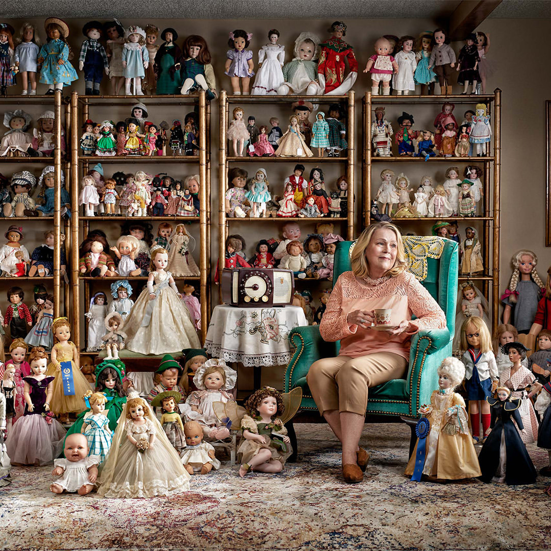 Mature woman sits surrounded by a large doll collection on shelves and floor, holding a tea cup in an indoor setting with patterned rug.