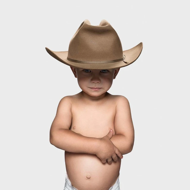 Toddler stands against a white backdrop with arms crossed, wearing a cowboy hat and a diaper.