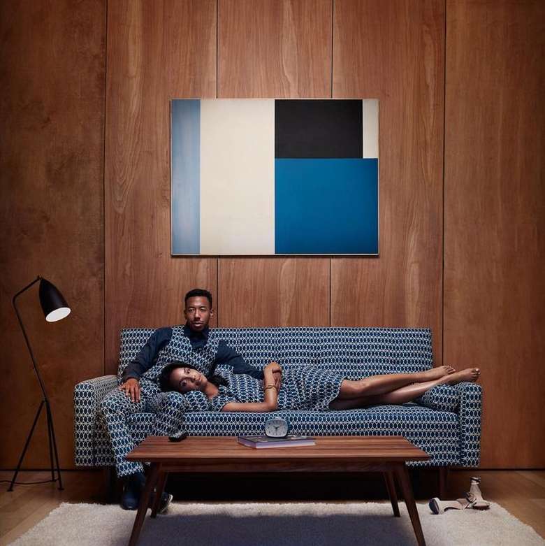 A stylish couple lounges on a patterned couch in a wood-paneled room, complemented by modern art and a sleek floor lamp.