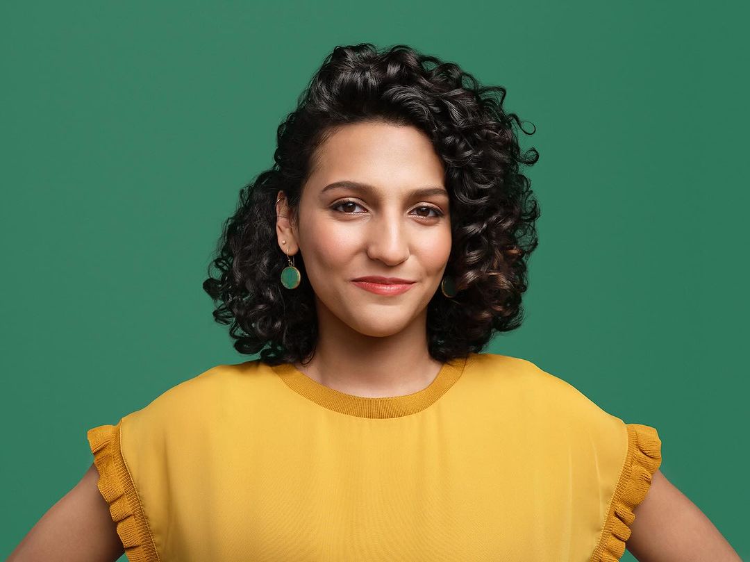 Smiling woman with curly hair poses in front of a solid green background, wearing a mustard yellow top and green earrings in a portrait.