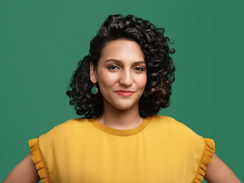 Smiling woman with curly hair poses in front of a solid green background, wearing a mustard yellow top and green earrings in a portrait.