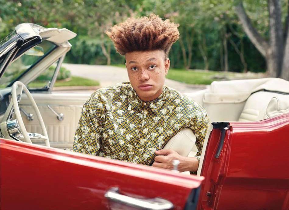 Adult male with curly hair wearing a patterned shirt inside a red convertible car outdoors surrounded by green trees in daylight.
