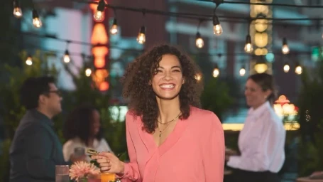 Smiling woman in a pink blouse holding a glass outdoors under string lights at night, photographed by Randal Ford.