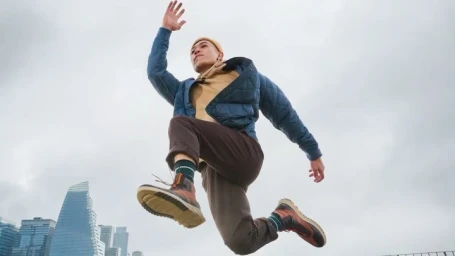 Man wearing a blue jacket, brown pants, and boots jumping in mid-air against an overcast sky with distant skyscrapers