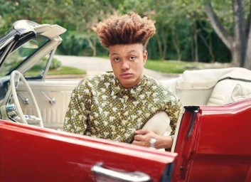 Young man with tall curly hair wearing an olive green patterned shirt seated inside a red convertible car.