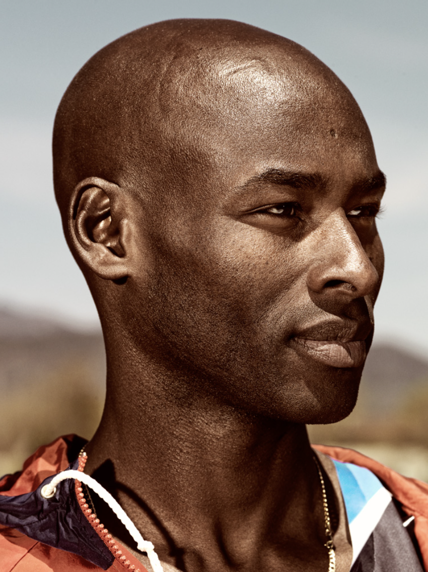 Close-up portrait of a bald African American man looking to the right, wearing an orange jacket against a blurred background.