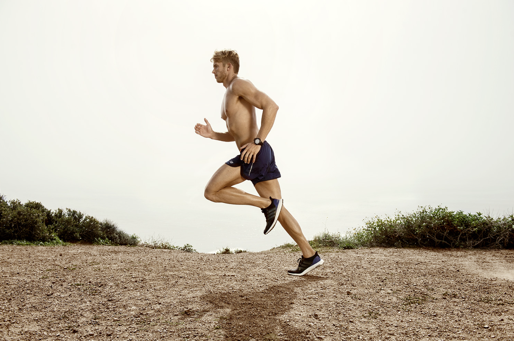 Athletic blond man with bare torso is running outdoors on a dirt path. He is wearing dark athletic shorts and black running shoes.