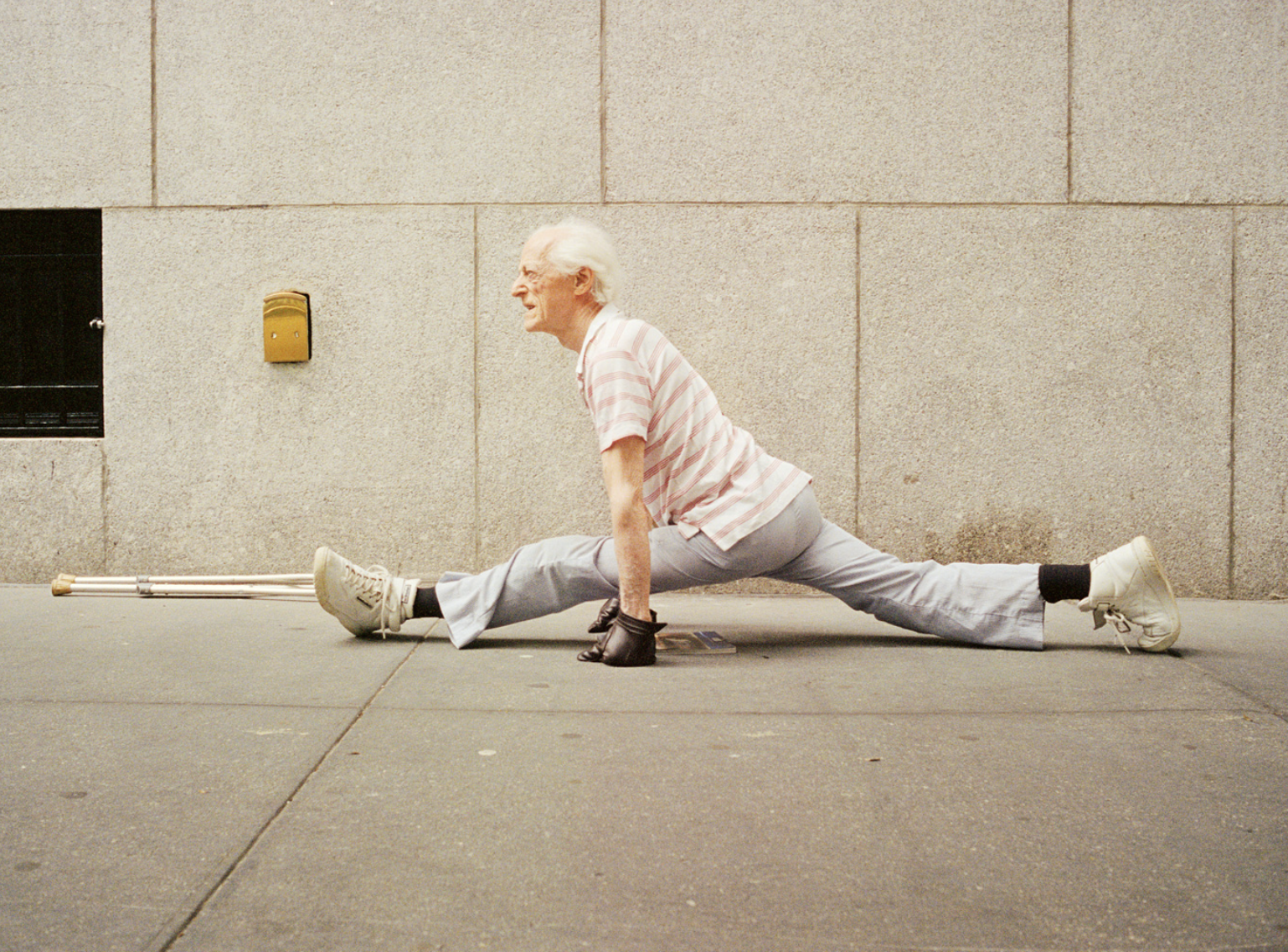 Elderly man performing splits on sidewalk outdoors, wearing casual clothing, with crutches lying beside him against a concrete wall.