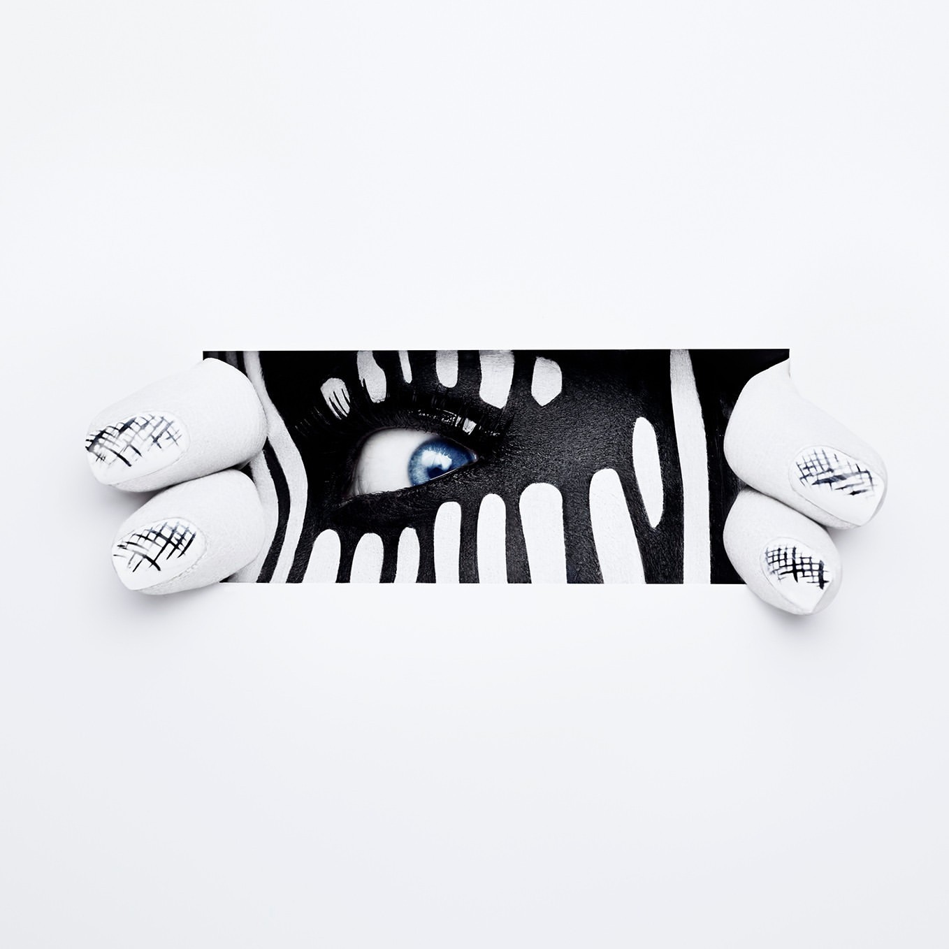 Close-up of a person's eye with artistic black and white zebra-like face paint, peeking through a white paper frame, nail art visible.
