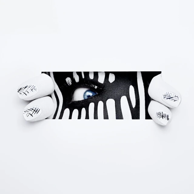 Close-up of a person's eye with artistic black and white zebra-like face paint, peeking through a white paper frame, nail art visible.