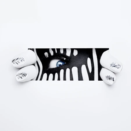 Close-up of a person's eye with artistic black and white zebra-like face paint, peeking through a white paper frame, nail art visible.