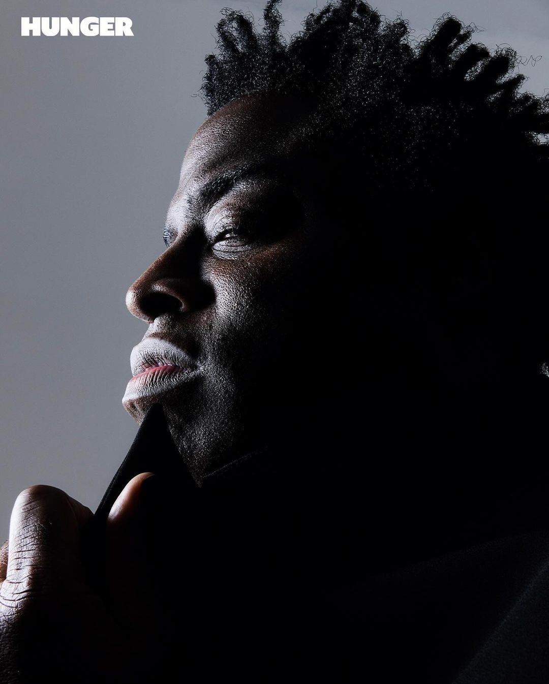 Portrait of a Black male model with high-fashion makeup looking left, against a grey background in a studio with side lighting. 