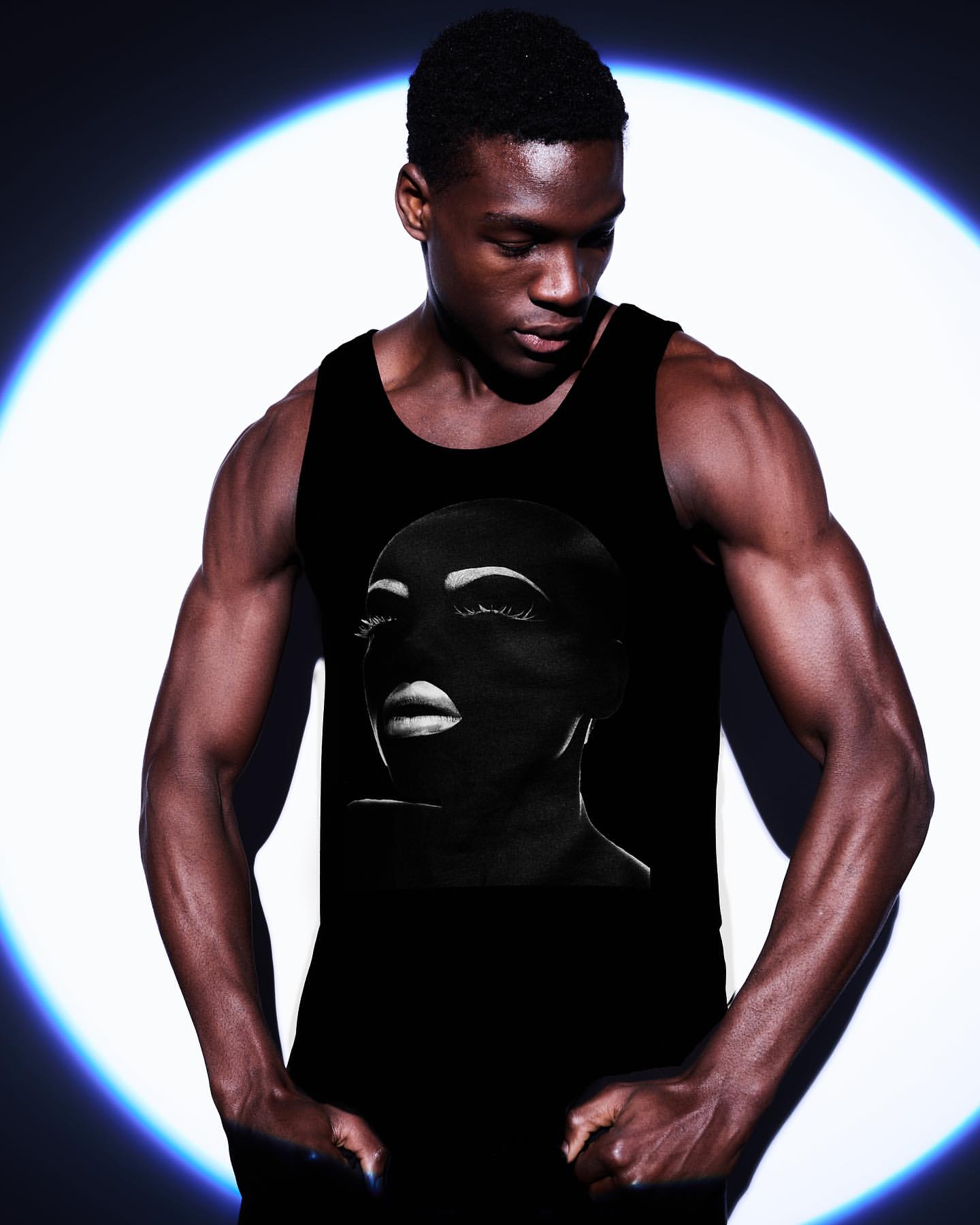 An athletic Black male model with strong biceps and a black tank top featuring a black and white portrait. The background is dark with bright lighting.