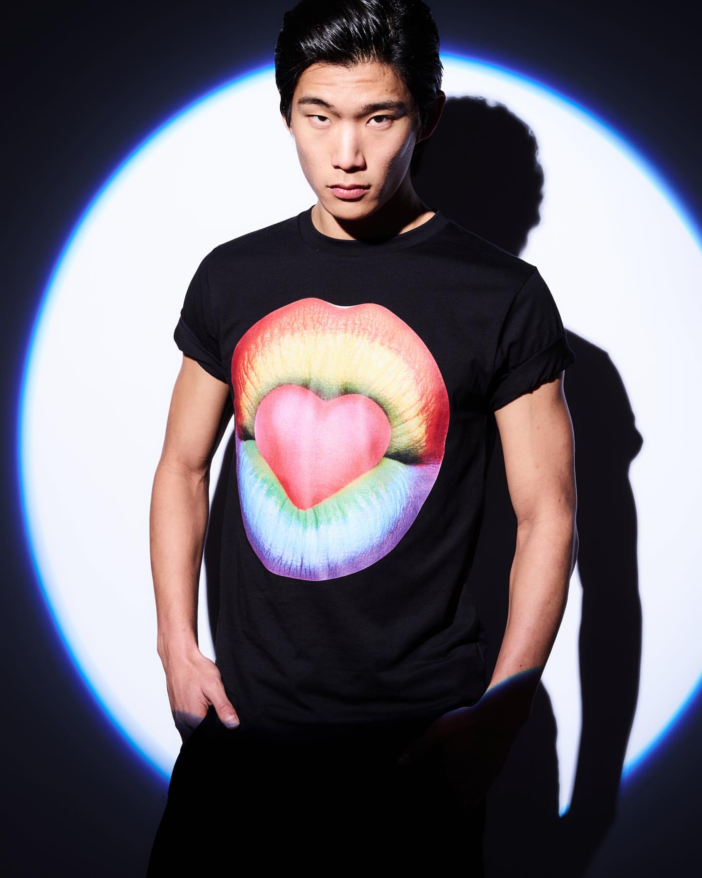 East Asian man wearing a black t-shirt with a rainbow lips graphic, posed against a circular blue spotlight backdrop.