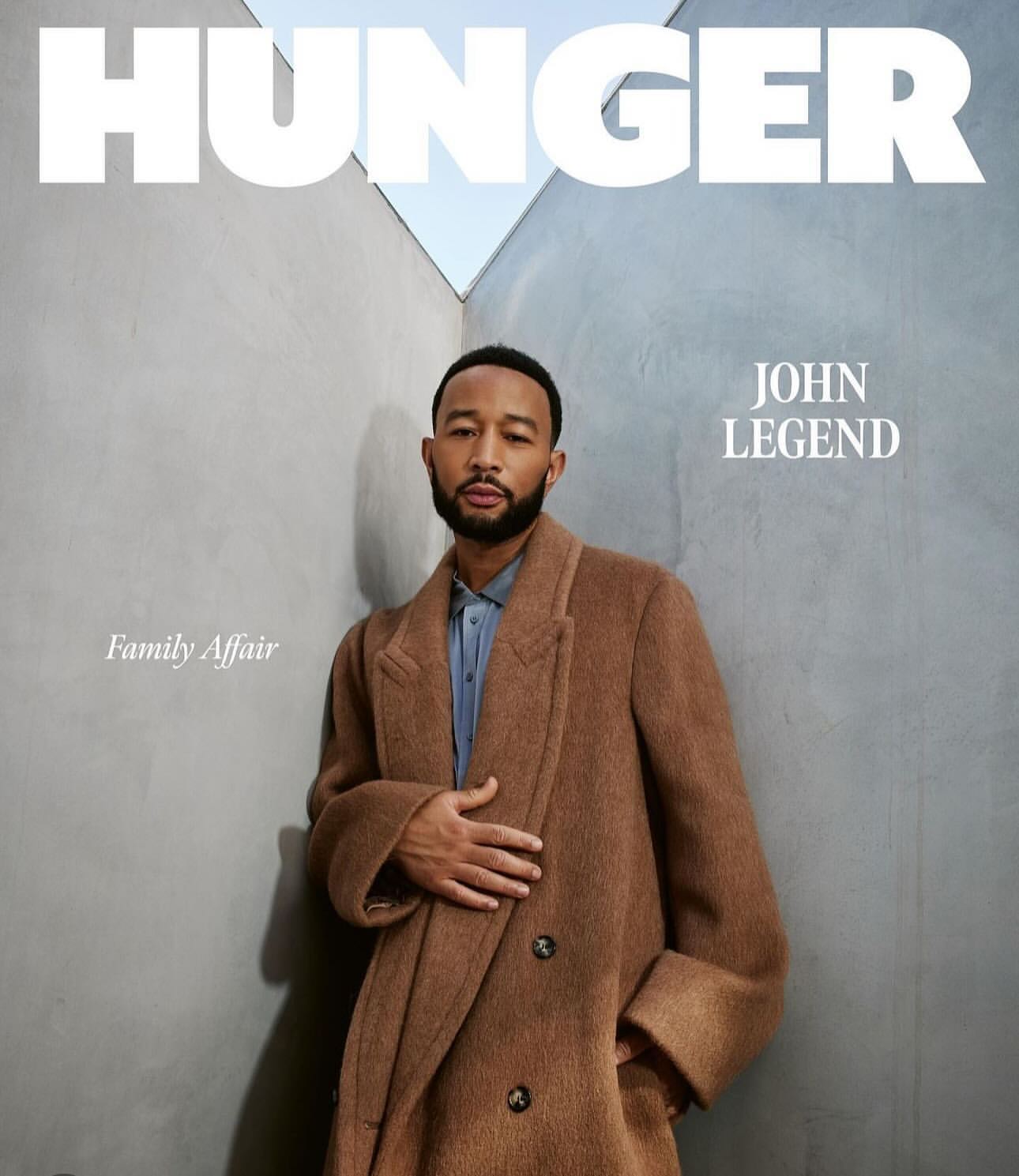 John Legend poses in brown coat for Hunger magazine cover against a concrete wall, highlighting fashion and celebrity portrait photography.