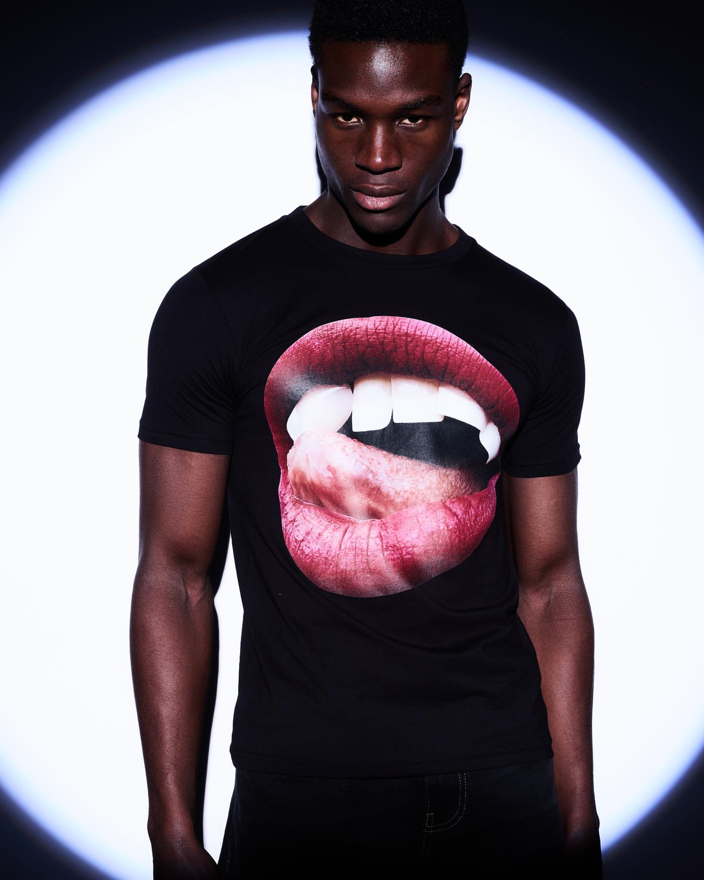 Fashion shot of a Black adult male wearing a black t-shirt with a mouth graphic, posing in front of a bright, round light source.