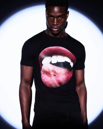 Fashion shot of a Black adult male wearing a black t-shirt with a mouth graphic, posing in front of a bright, round light source.