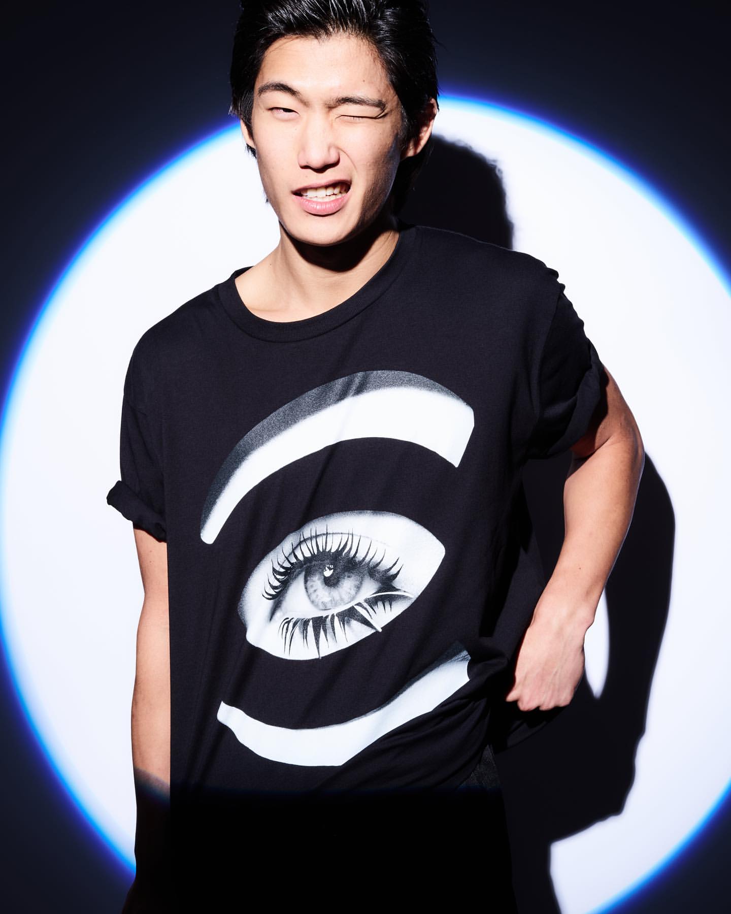 A young adult male with black hair poses indoors wearing a black t-shirt with a stylized eye graphic against a spotlit background.